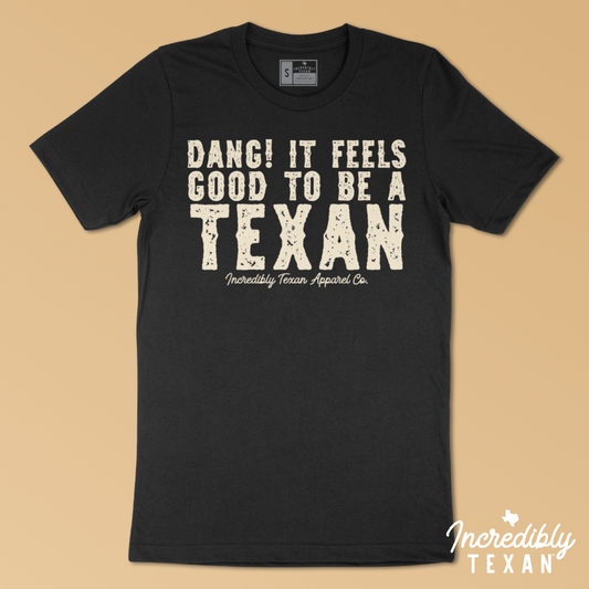 A black, short sleeve shirt with a light beige print on the front that reads "DANG! IT FEELS GOOD TO BE A TEXAN - Incredibly Texan Apparel Co." in an Old West font.